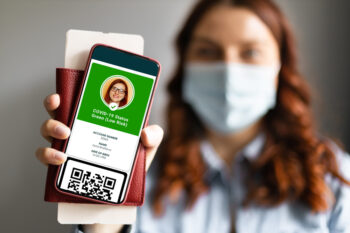 Young traveler woman in protective medical mask shows health passport of vaccination certification on phone at airport or train platform, to certify that have been vaccinated of coronavirus covid-19
