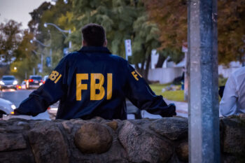 Male FBI agent wearing dark blue coat with FBI logo looking down the street with cars in the dusk seen from behind.