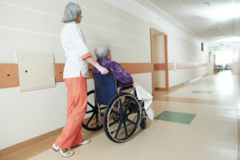 female nurse carer and aged elderly patient woman in wheelchair at clinic hallway