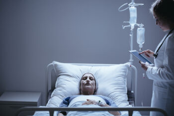 Doctor with tablet next to bed with dying elderly woman with cancer in the clinic